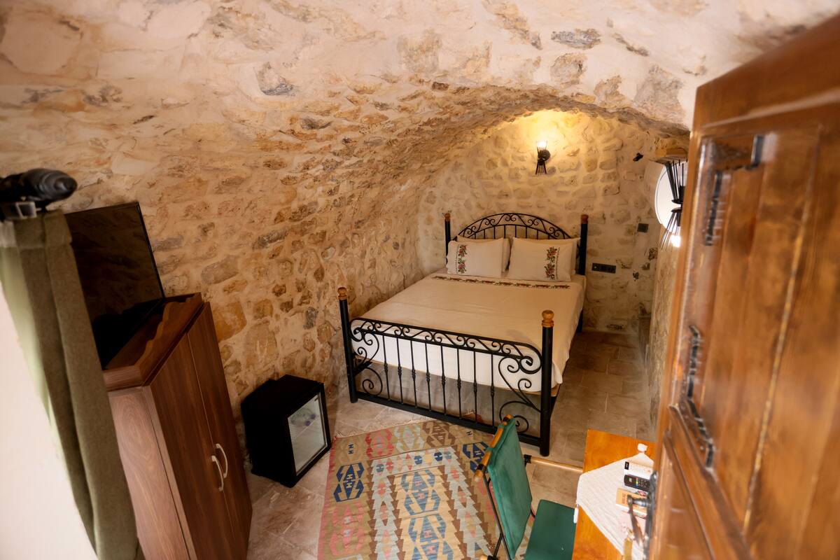 A stone-walled bedroom with a metal bed frame, a small bedside table, and a green chair.