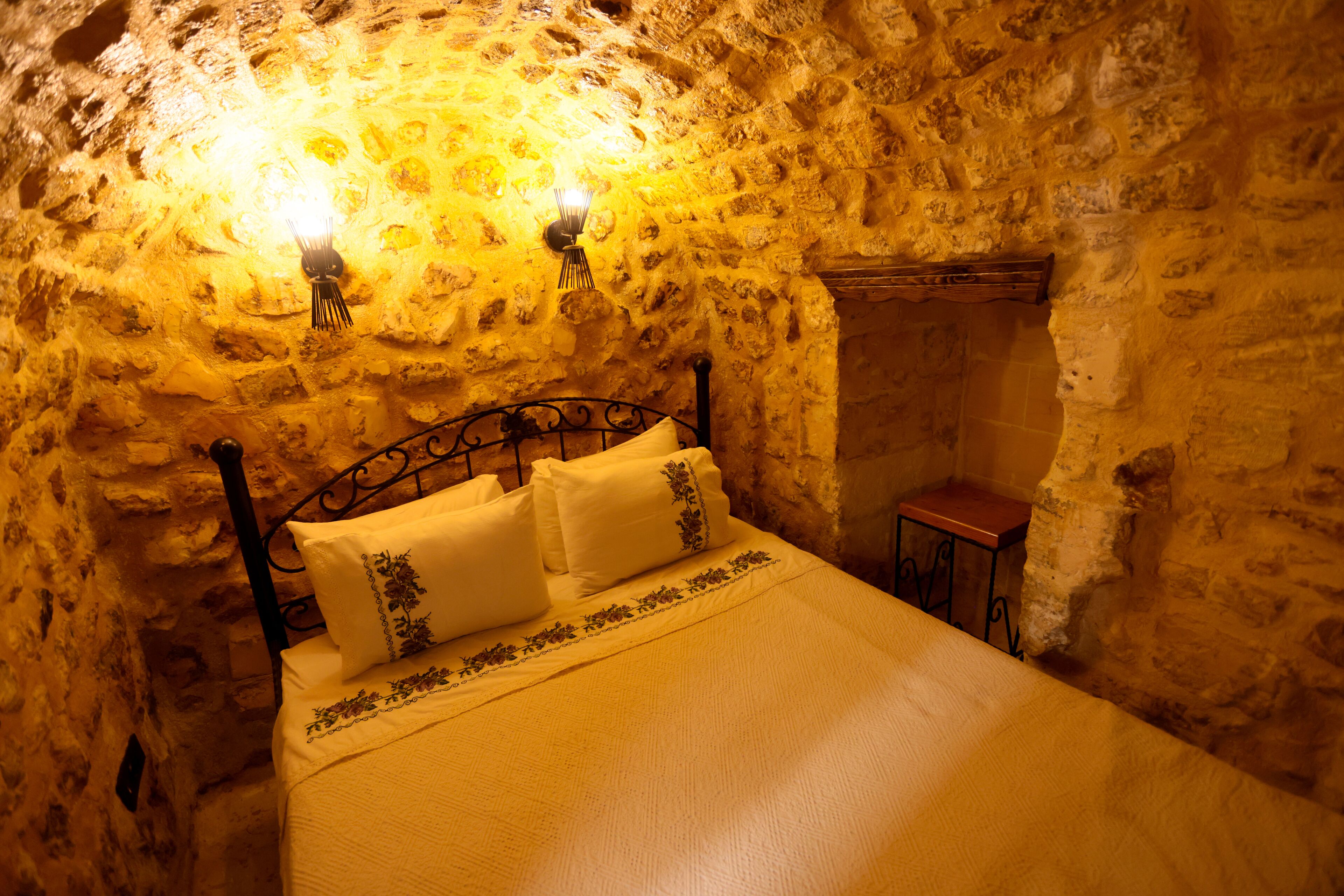 A stone-walled room with a bed, two wall-mounted lights, and a small table.