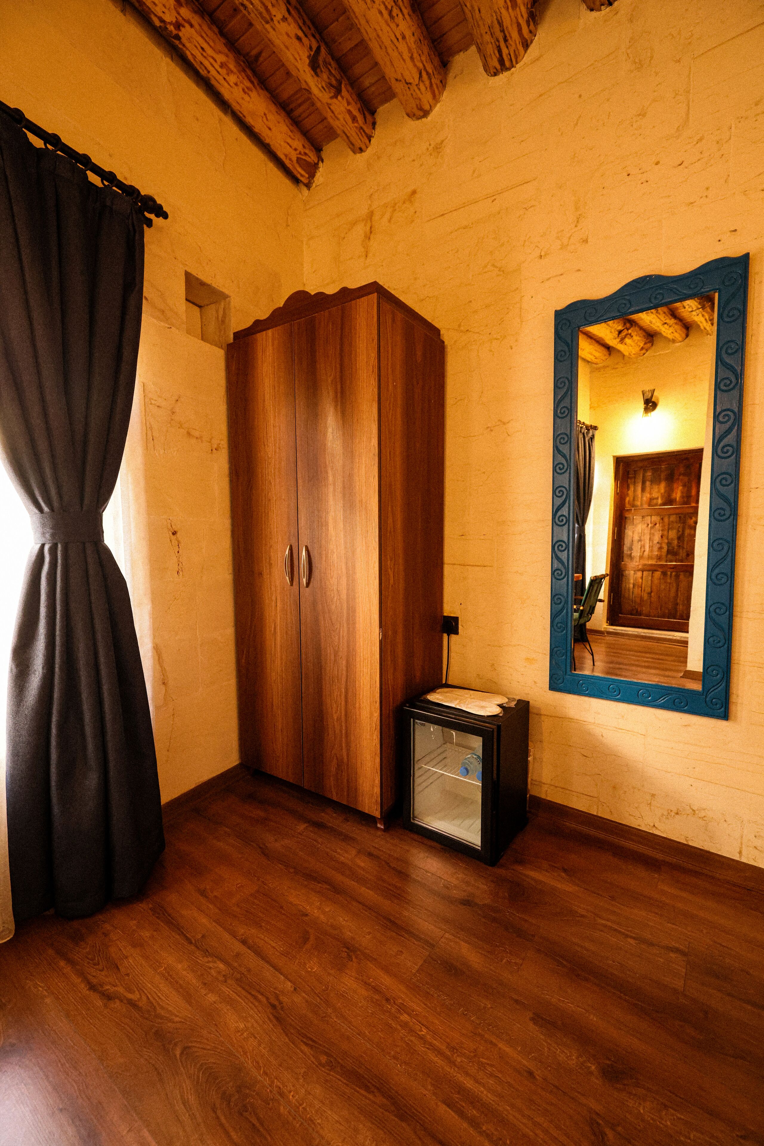 A room with wooden flooring, a wooden wardrobe, a mirror with a decorative frame, and a small glass cabinet.