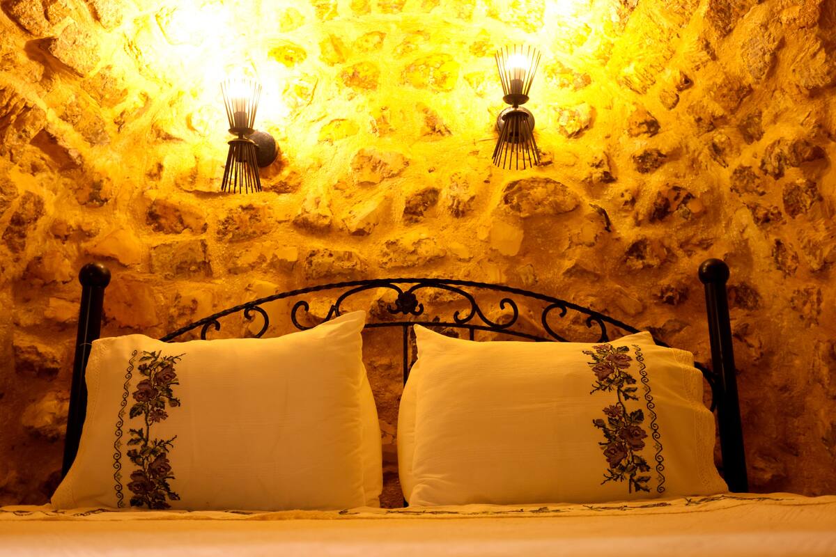 A bed with a wrought iron headboard, two pillows with floral embroidery, and a stone wall with two wall-mounted light fixtures.