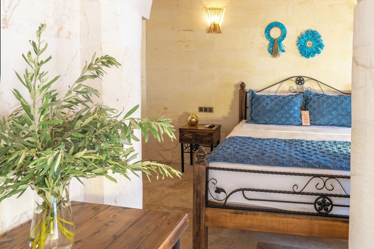 A bedroom with a wooden bed, a nightstand, a vase with greenery, and a stone wall.