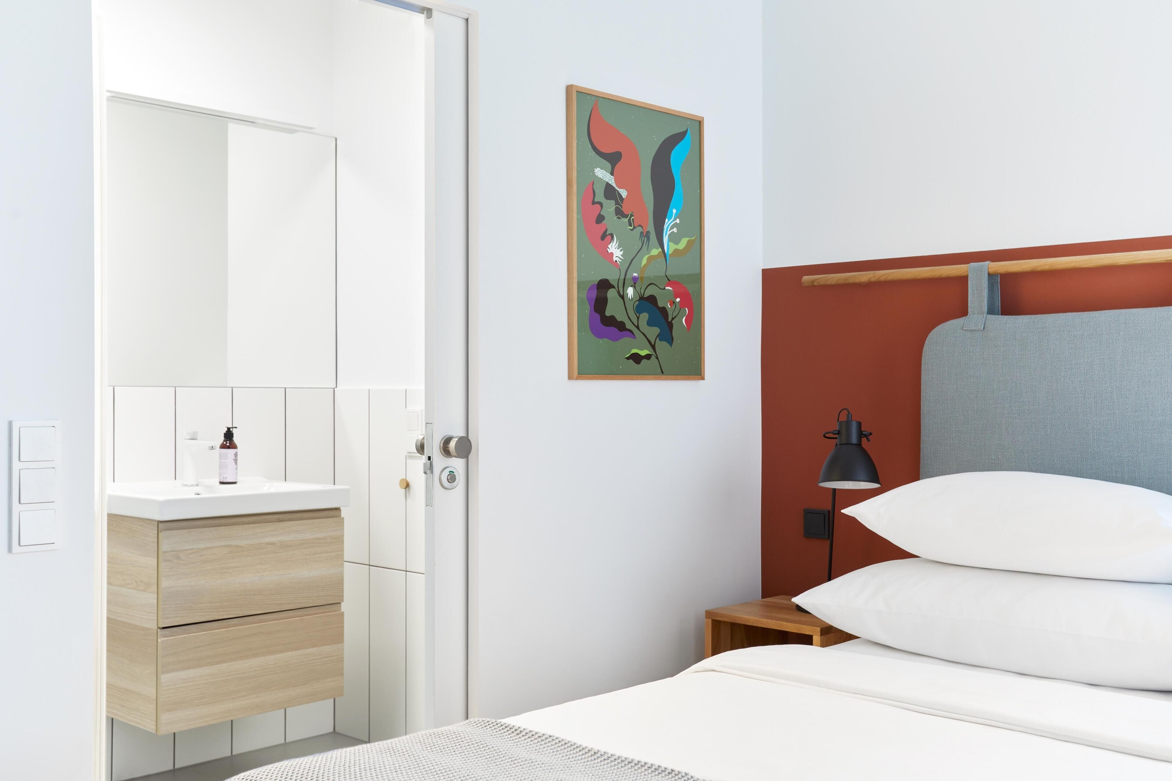 Una habitación de hotel moderna con una cama blanca, una mesita de noche de madera, una mesita de noche con una lámpara, una pared blanca, un cuadro abstracto enmarcado y una puerta blanca.