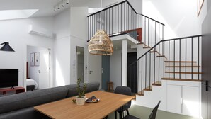 A modern living room with a wooden dining table, grey sofa, and black chairs. A large woven light fixture hangs from the ceiling, and a staircase with black railings leads to an upper level. - Plantonia (Kraków)