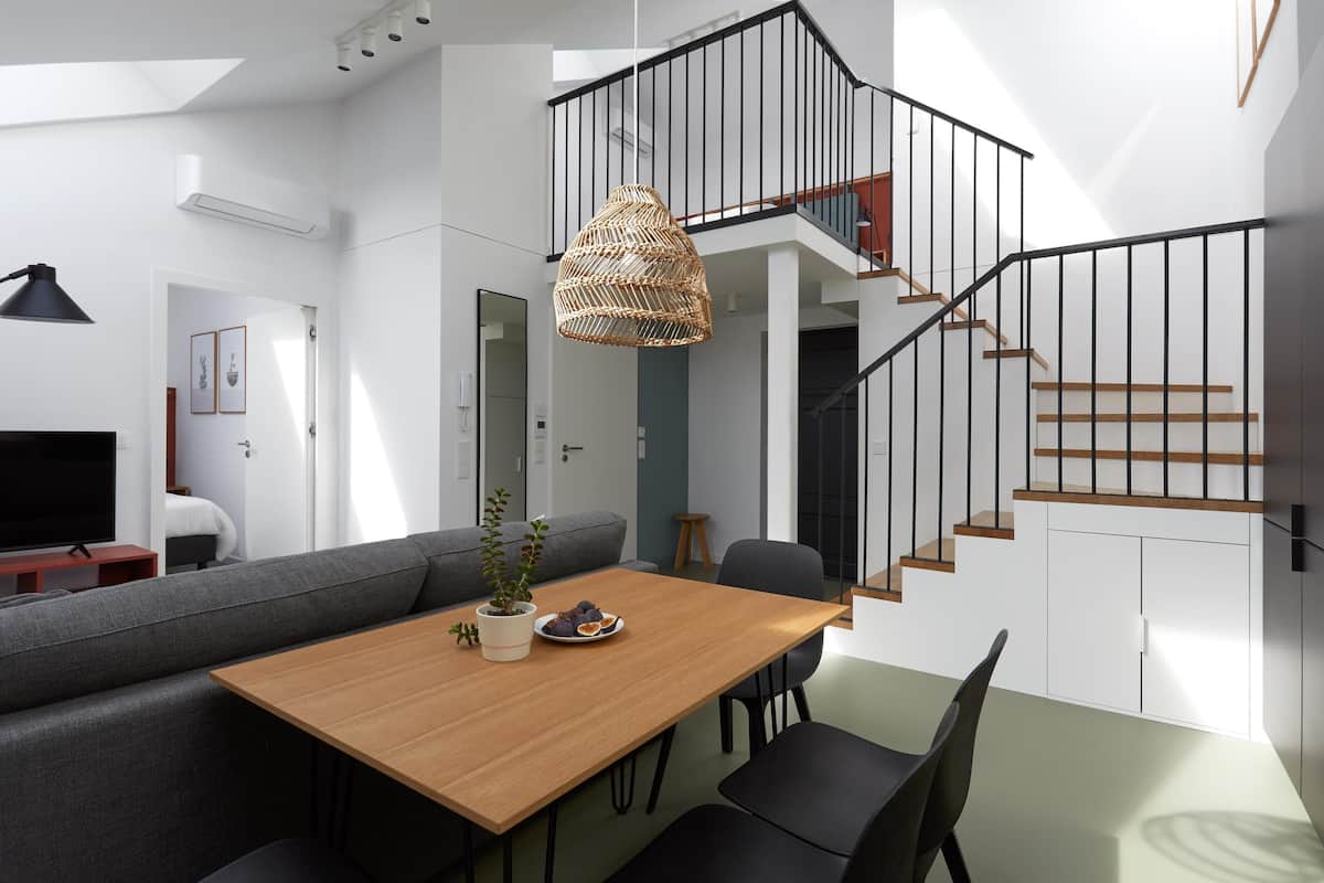 A modern living room with a wooden dining table, grey sofa, and black chairs. A large woven light fixture hangs from the ceiling, and a staircase with black railings leads to an upper level.