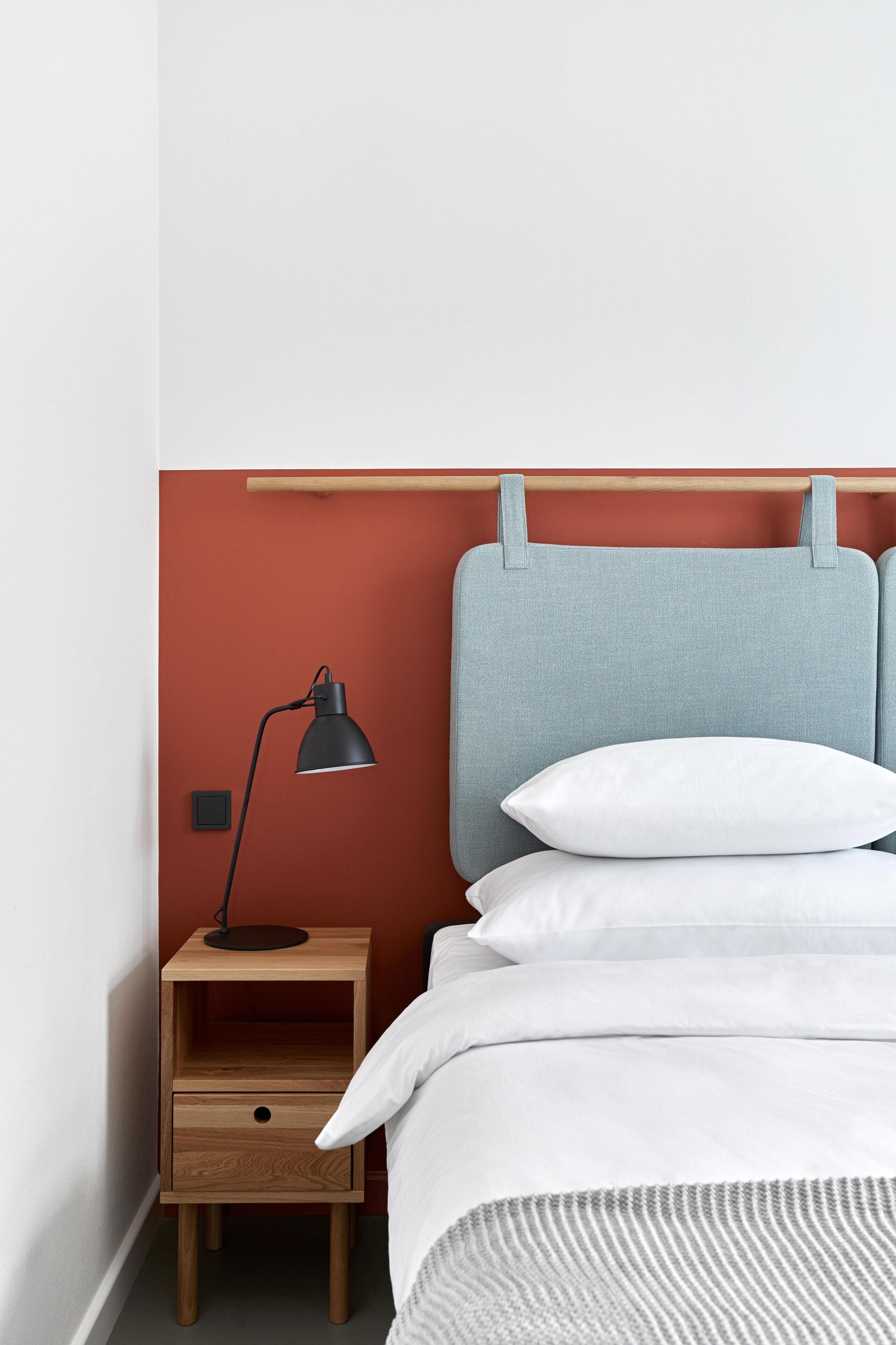 Un lit bien fait, avec du linge de lit blanc et une couverture rayée grise, une petite table de chevet en bois avec une lampe noire, et un mur rouge.