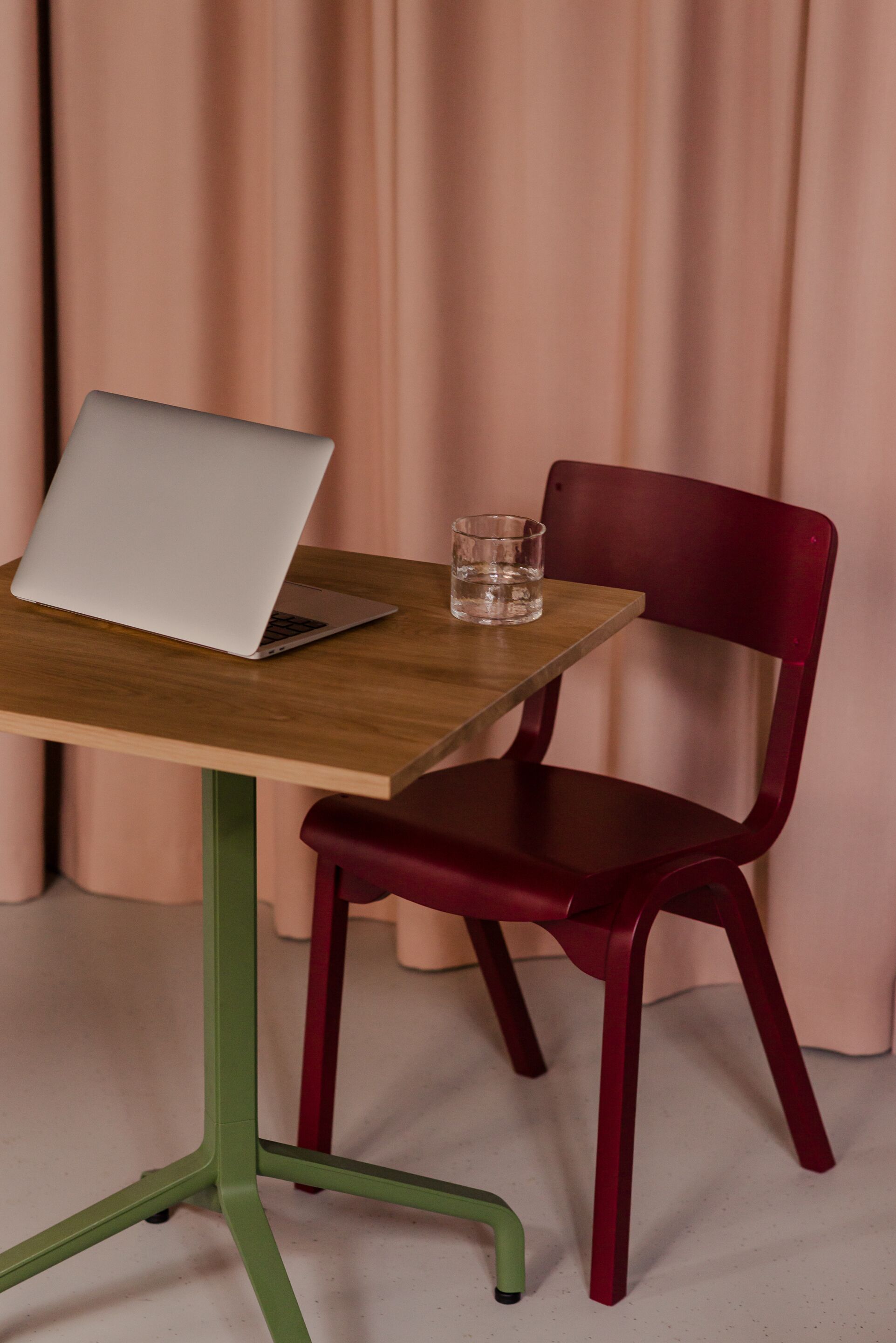 A wooden table with a laptop and a glass of water, a red chair, and a green metal chair leg.