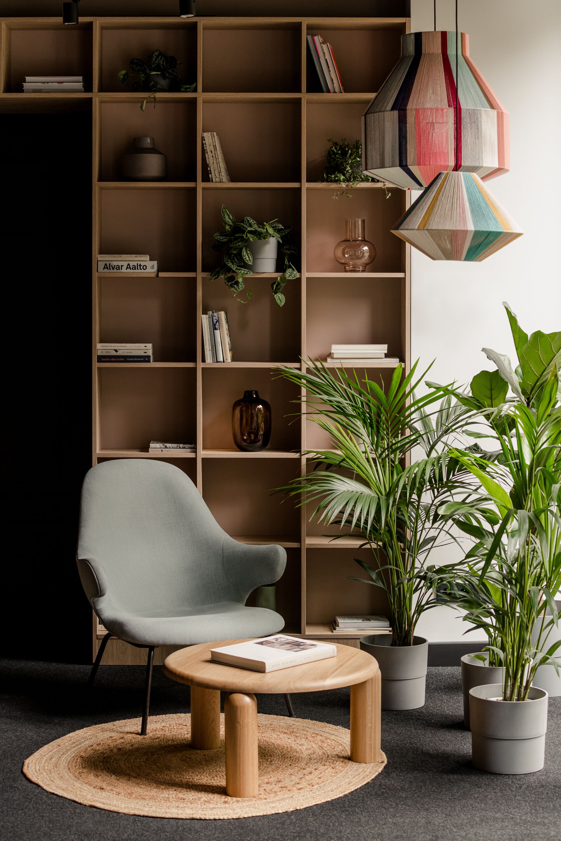 Una sala de estar moderna con un sillón gris, una mesa redonda de madera, una estantería llena de libros y objetos decorativos, y varias plantas en macetas.