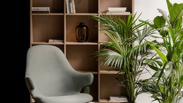 Una sala de estar moderna con un sillón gris, una mesa redonda de madera, una estantería llena de libros y objetos decorativos, y varias plantas en macetas.