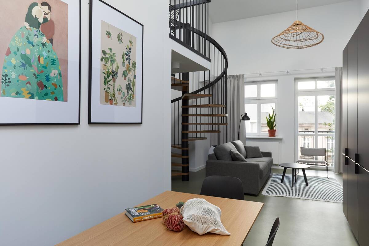 A modern living room with a spiral staircase, a grey sofa, a dining table with a book and a woven bag, and two framed artworks on the wall.