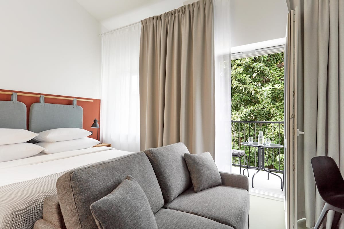 A hotel room with a bed, grey sofa, and a balcony with a table and chairs.