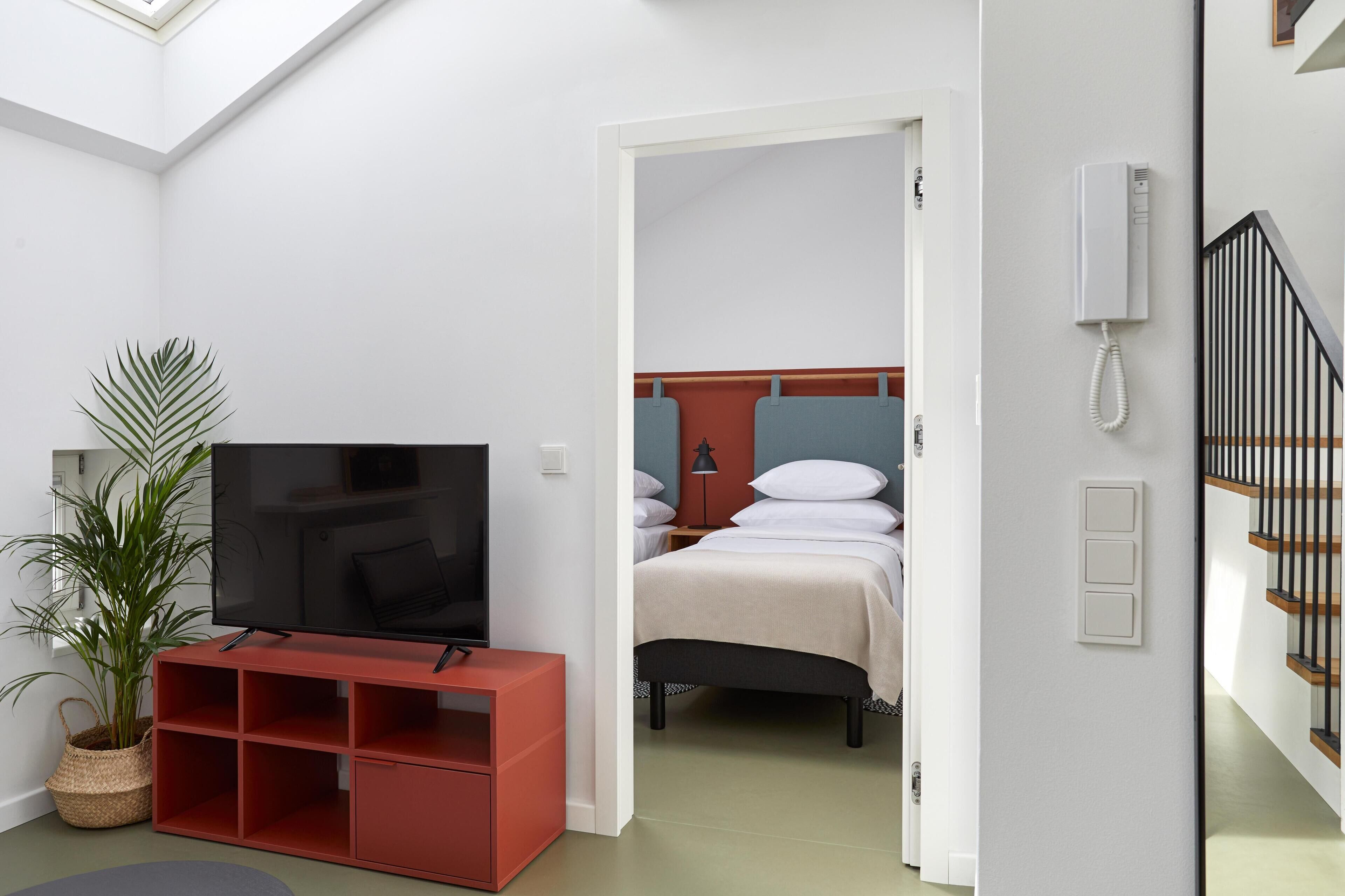 Een hotelkamer met een televisie, een plant, een rode opbergunit, een bed met witte beddengoed en een trap.
