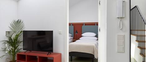 Habitación de hotel con televisor, planta, armario rojo, cama con ropa de cama blanca y una escalera.