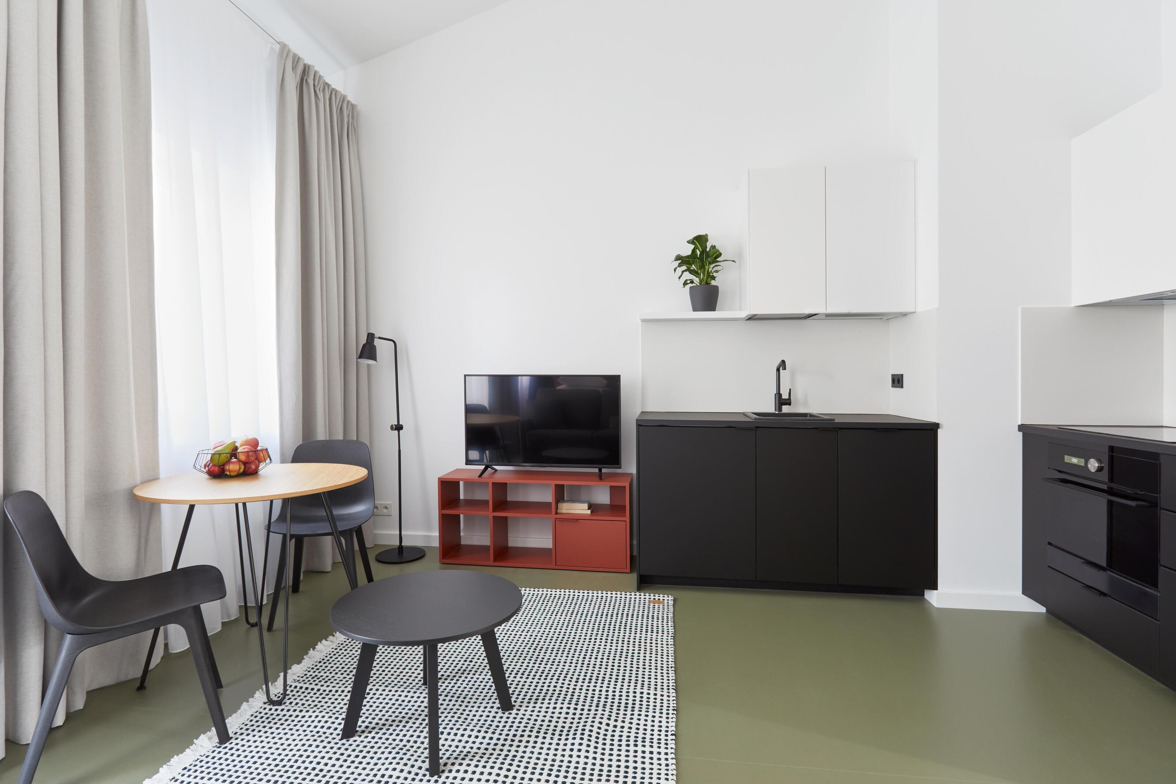 Ett modernt vardagsrum med en matplats, en platt-TV, ett litet kök med diskho och en krukväxt.