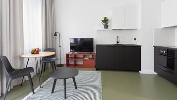 Ett modernt vardagsrum med en matplats, en platt-TV, ett litet kök med diskho och en krukväxt.
