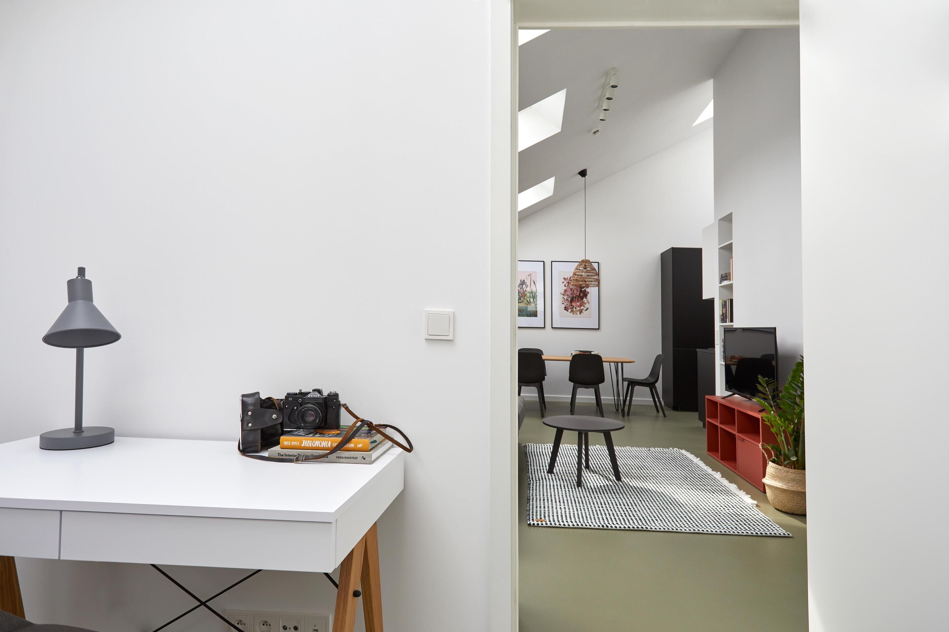 Een moderne woonruimte met een wit bureau, een camera en boeken, die uitkomt op een eethoek met stoelen en een tafel.