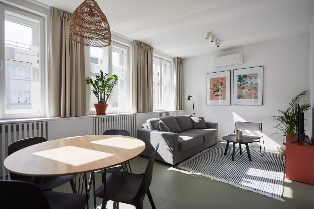 A modern living room with a grey sofa, a round wooden table with chairs, a black floor lamp, and a large window with curtains.