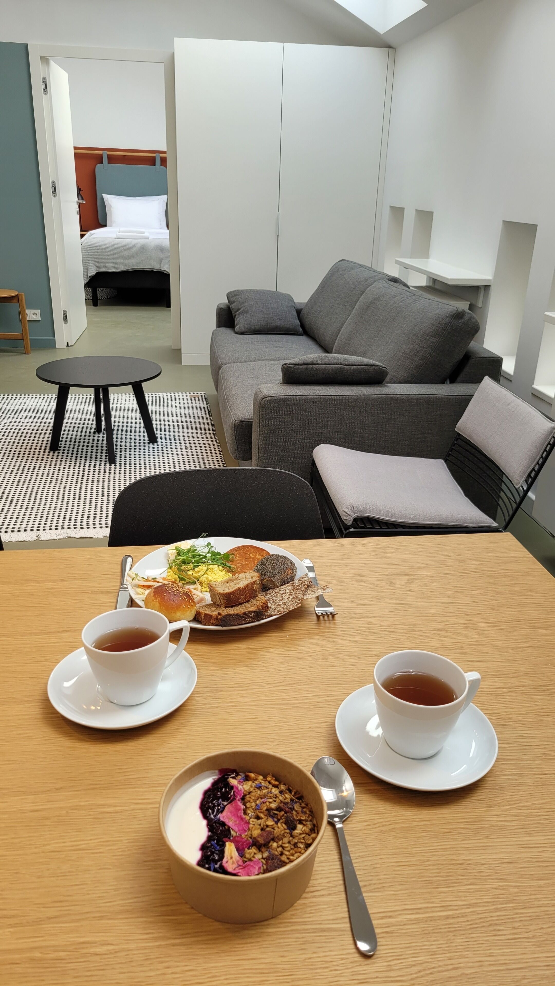 A modern hotel room with a grey sofa, a small round table with breakfast items, and a bed in the background.