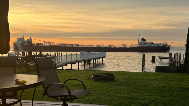 Een terras met buitenmeubilair, uitkijkend op een waterkant met een steiger en een groot schip in de verte.