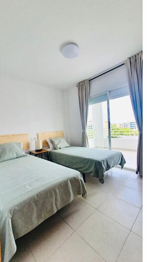 Two single beds with wooden headboards and green bedspreads, a large window with curtains, and a tiled floor.