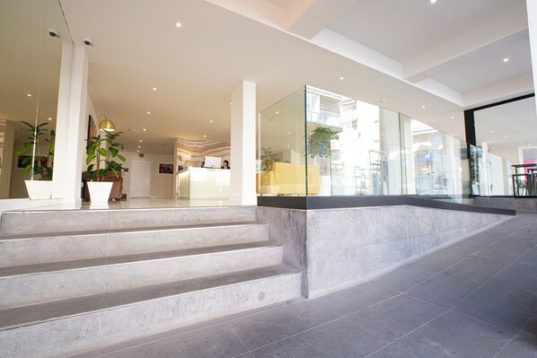 Un hall d’hôtel moderne avec un comptoir d’accueil, un escalier et un mur de verre donnant sur la rue.