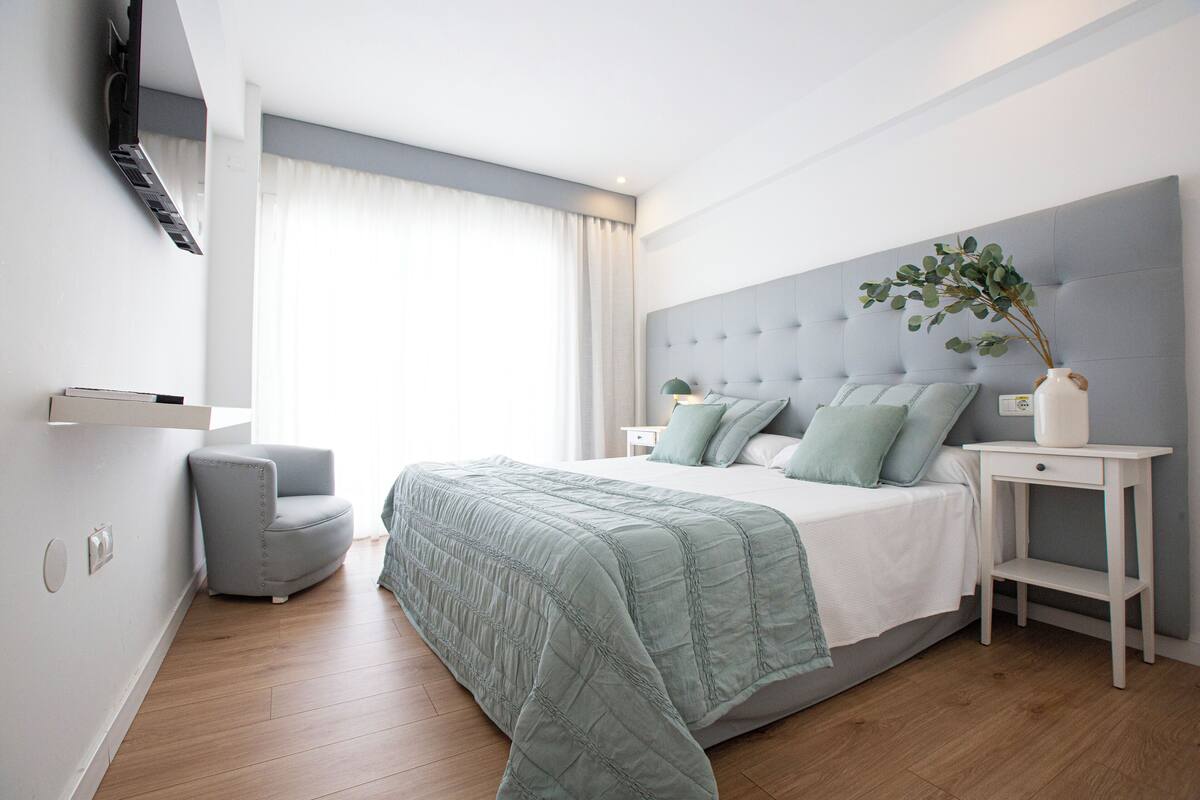 A modern bedroom with a large bed, a grey upholstered headboard, a bedside table with a lamp, and a small grey armchair.