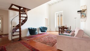 Ein geräumiges Wohnzimmer mit Wendeltreppe, einem weißen Sofa, zwei blauen Sesseln, einem Esstisch mit Stühlen und einem großen Spiegel an der Wand.