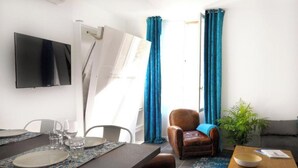 Een woonkamer met een eettafel gedekt voor twee personen, een bruine leren fauteuil, een aan de muur bevestigde televisie en een potplant.