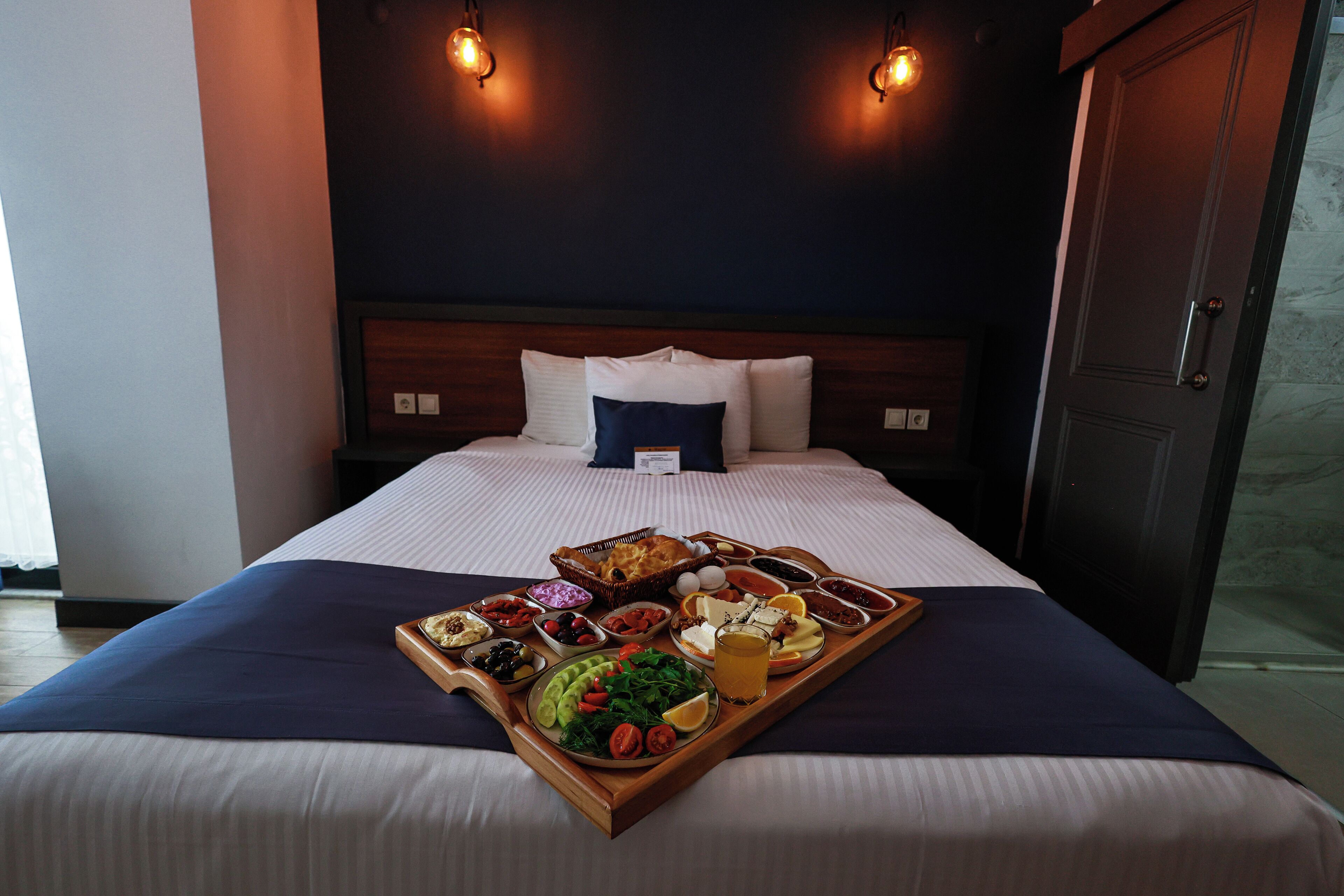 A neatly made bed with a tray of breakfast items, including eggs, toast, and fruit, in a hotel room with a dark headboard and two pendant lights.