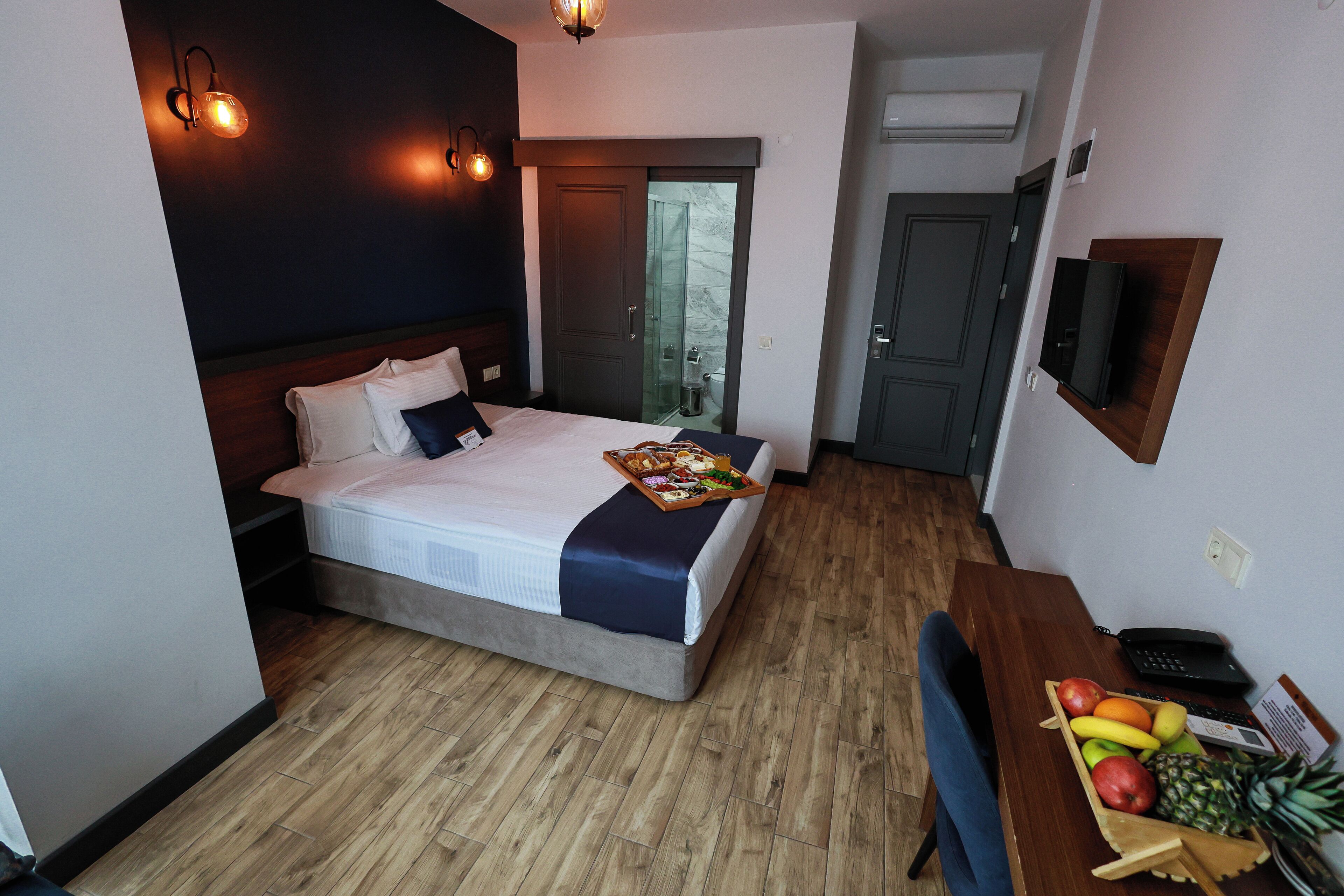 Una habitación de hotel con una cama, mesita de noche con frutas, un escritorio con teléfono y un televisor en la pared.