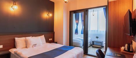 A hotel room with a large bed, a desk with a chair, a TV, and a sliding glass door leading to a balcony.