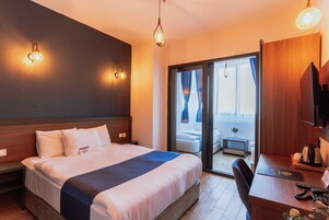 A hotel room with a large bed, a desk with a chair, a TV, and a sliding glass door leading to a balcony.