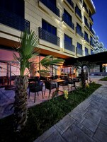 An outdoor seating area with tables and chairs, surrounded by palm trees and a building with balconies.