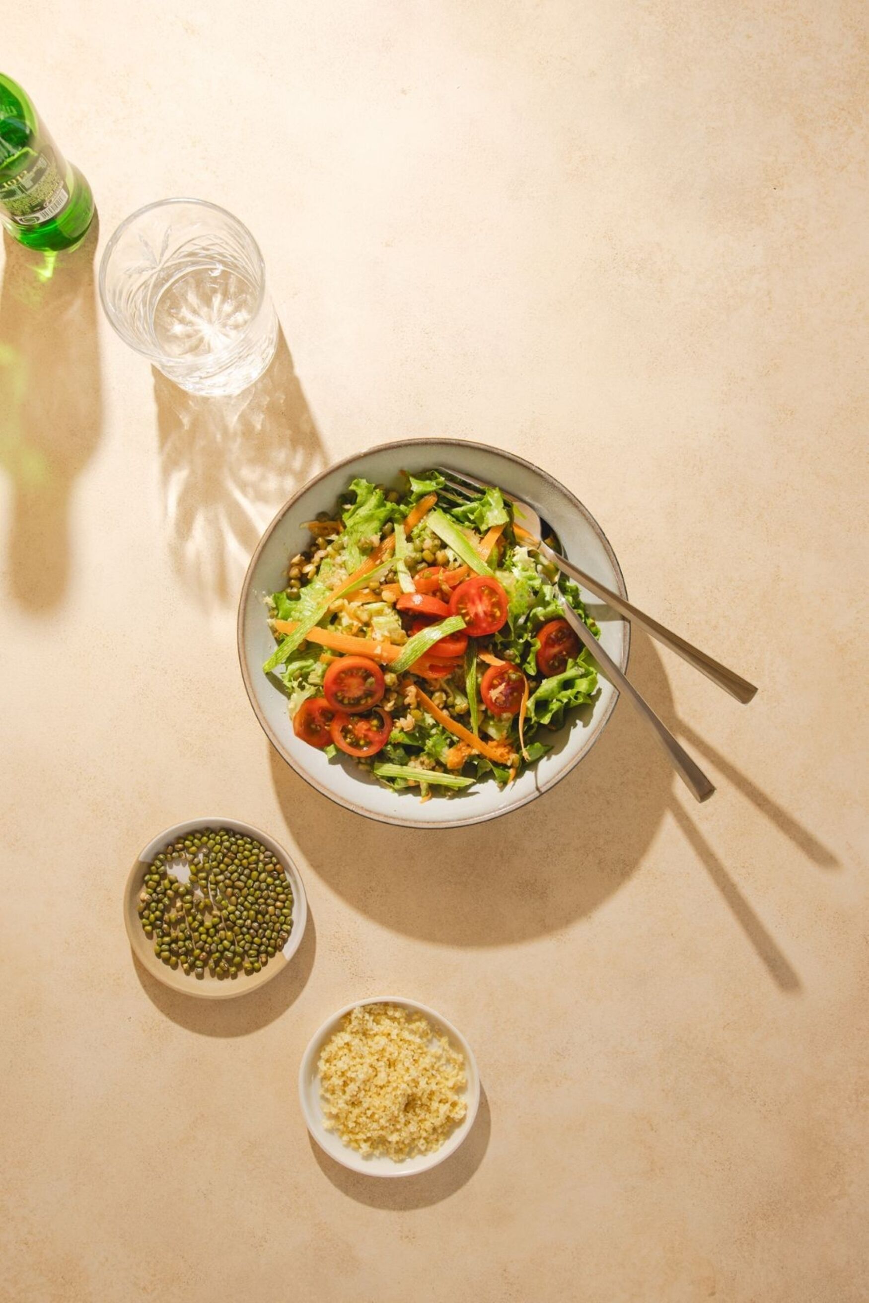 A bowl of salad with a fork, two small bowls of seeds, and a glass of water on a beige surface.