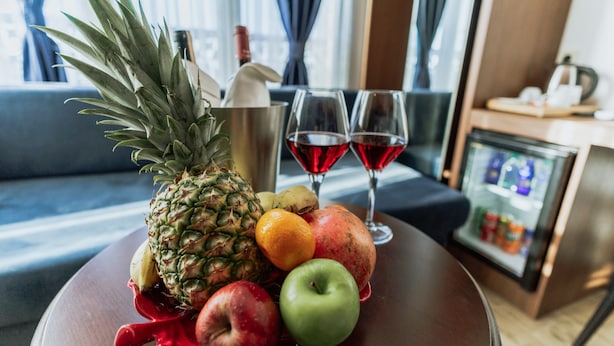 Una mesa con frutas, dos copas de vino tinto, un minibar y un sofá al fondo.