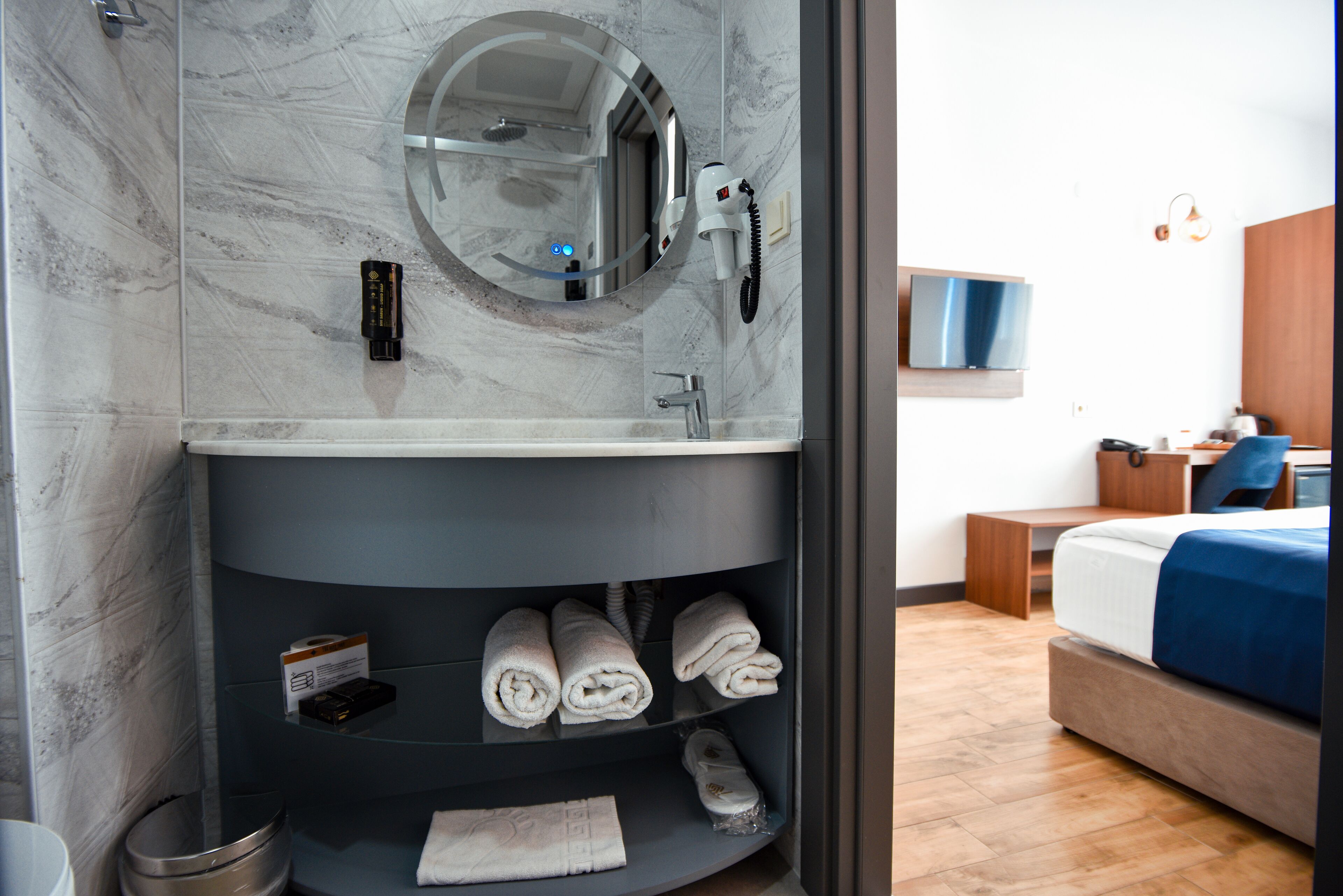 A hotel bathroom with a marble wall, a round mirror, a hairdryer, and a shelf with towels.