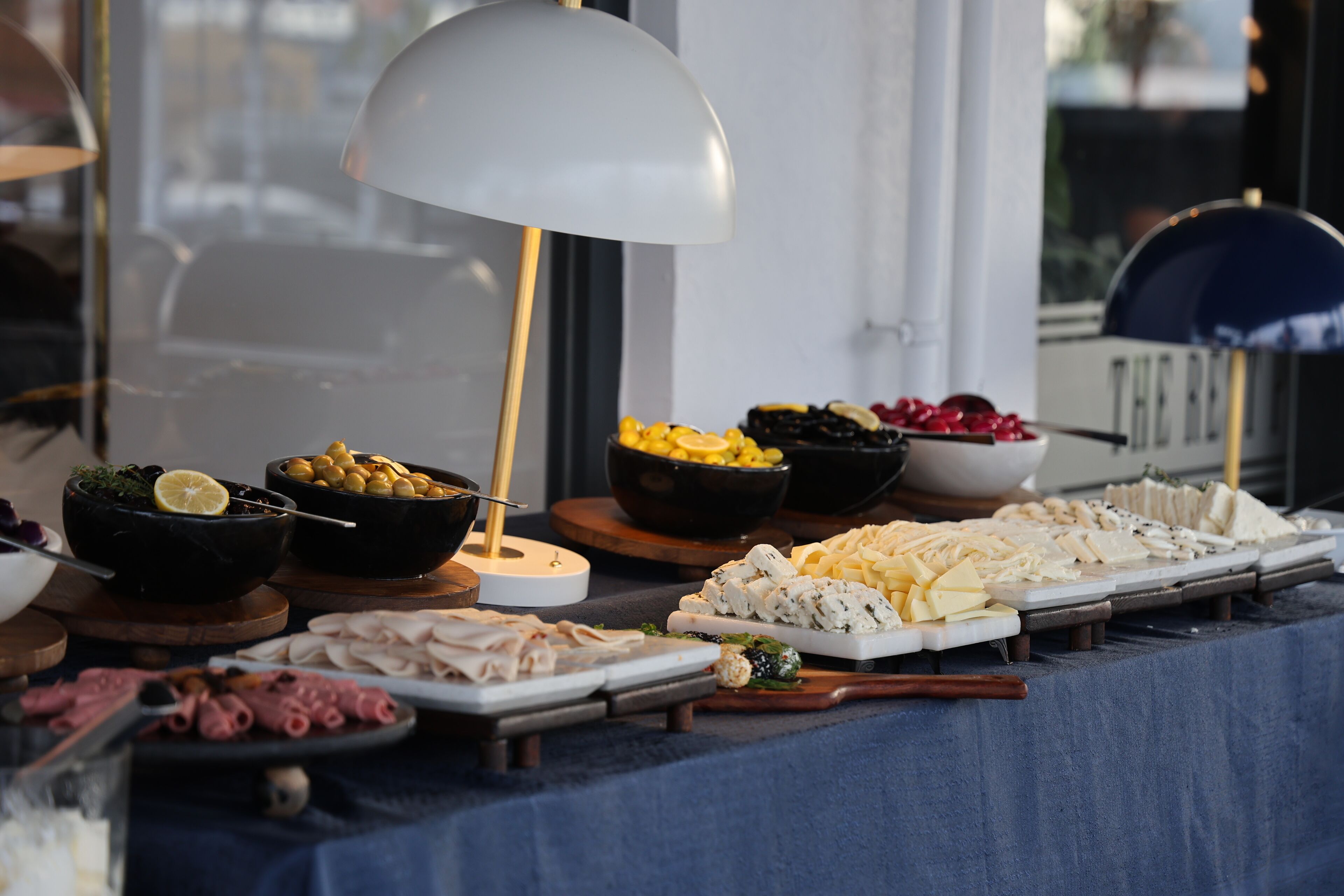 Una mesa de buffet con diversos alimentos, como quesos, carnes y aceitunas, bajo una lámpara blanca grande.