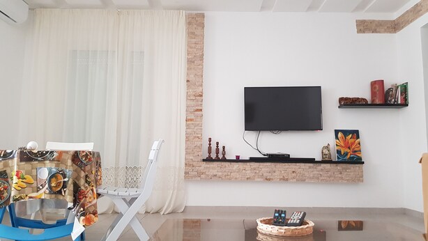 A modern living room with a flat-screen TV mounted on a brick wall, a dining area with a table set for two, and a decorative shelf with books and a clock.