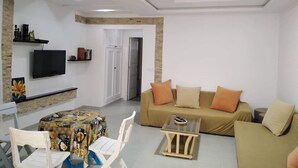 A living room with a sofa, a TV mounted on the wall, a table with chairs, and a door leading to another room.