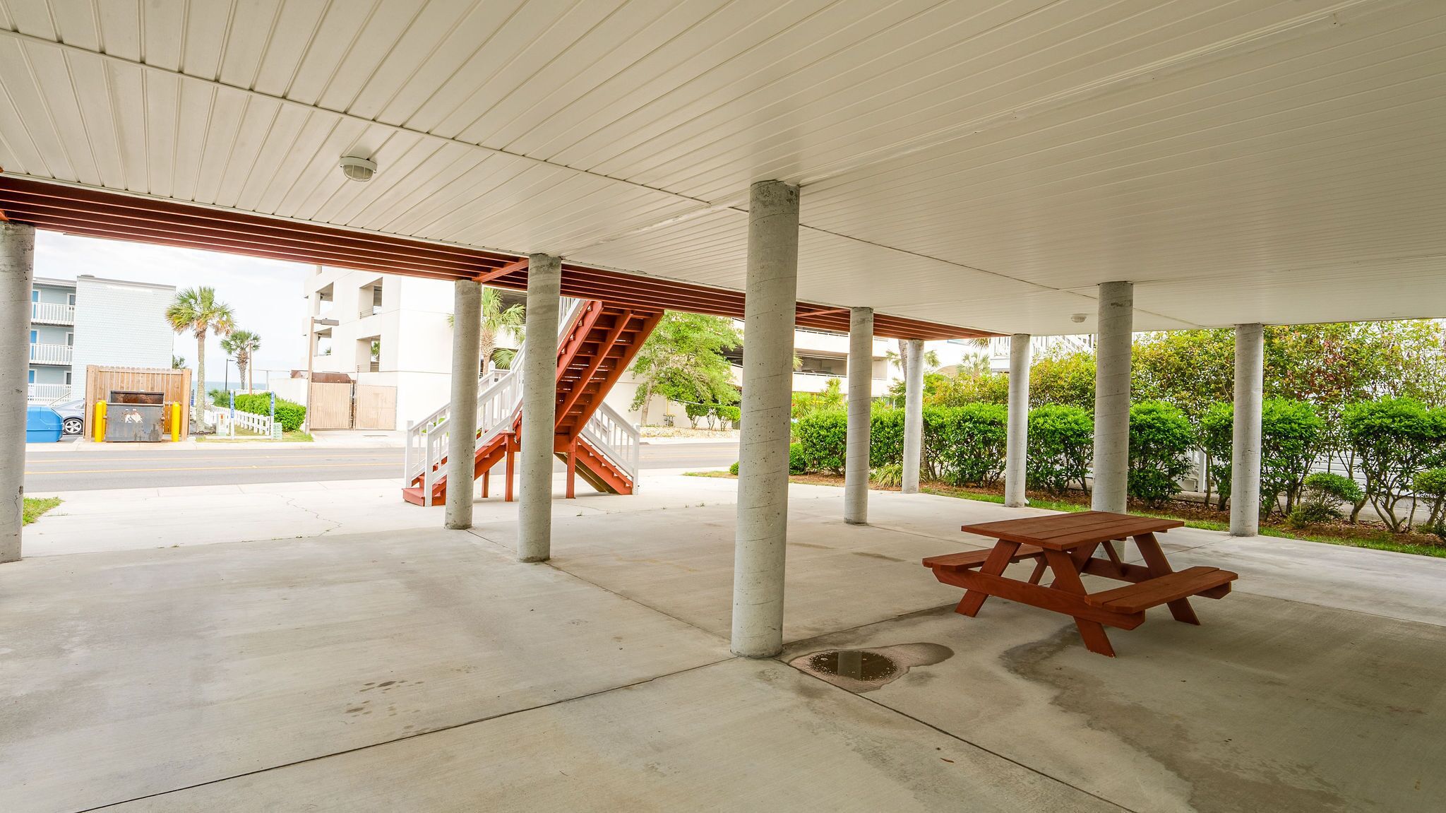 An outdoor covered area with concrete pillars, a wooden picnic table, and a staircase leading to an upper level.
