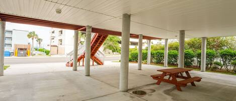 An outdoor covered area with concrete pillars, a wooden picnic table, and a staircase leading to an upper level.