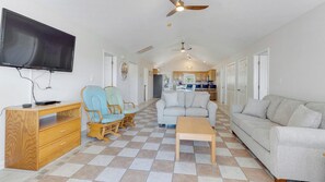 A spacious living area with a wooden desk, two armchairs, a sofa, and a television mounted on the wall. There is a ceiling fan, a kitchen in the background, and a tiled floor.