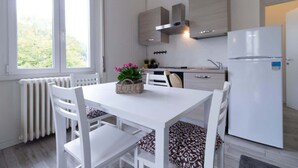 A modern kitchen with white cabinetry, a built-in stove, and a refrigerator. There is a dining table with chairs and a window providing natural light. - Casetta di Elie - Affitti Brevi Italia (Lecco)