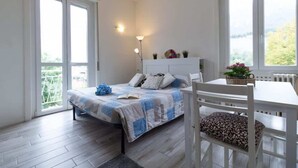 A bedroom with a bed, bedside table, chair, and a window with a view of greenery. - Casetta di Elie - Affitti Brevi Italia (Lecco)