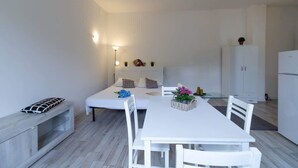 Een moderne, minimalistisch ingerichte kamer met een witte eettafel, witte stoelen, een bankje en een bed met een witte hoofdeinde. Achterin staat een klein kastje en een koelkast.