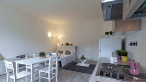 Een moderne keuken met witte kasten, een eettafel met stoelen, een bank en een kleine salontafel. Er staat een fornuis met pannen en een gootsteen.