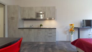 Een compacte keuken met grijze kastjes, een ingebouwde magnetron en een klein spoelgedeelte. Er staat een rode stoel en een bed met een rode deken.