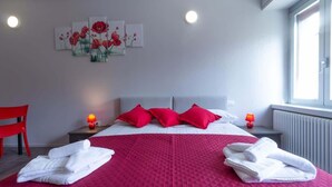 A hotel room with a bed, red pillows, white towels, a red quilted bedspread, a red chair, and a wall with a floral painting. - Appartamento Papavero - Affitti Brevi Italia (Lecco)