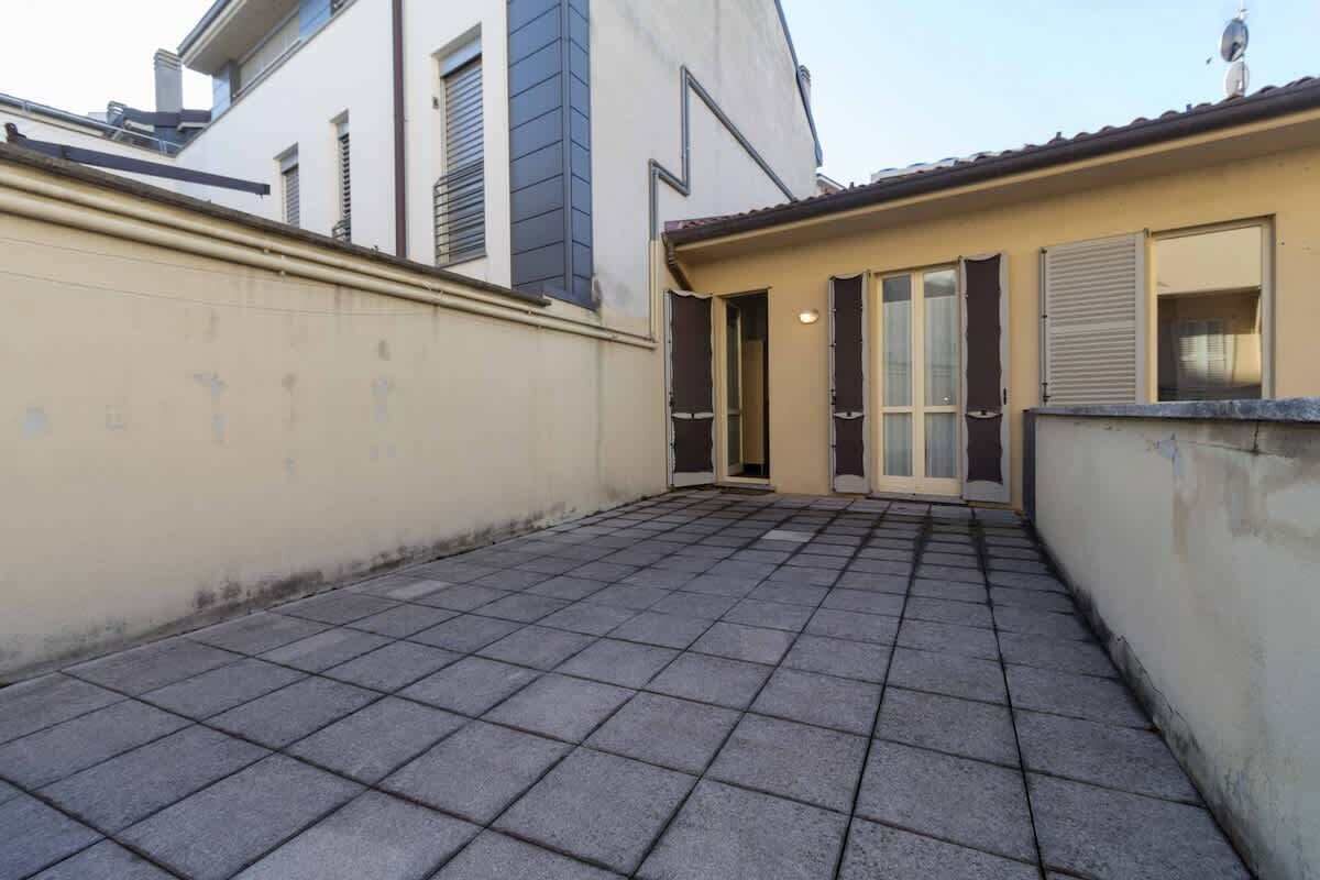 A courtyard with a tiled floor, a beige wall, and a building with a sloped roof and shutters.
