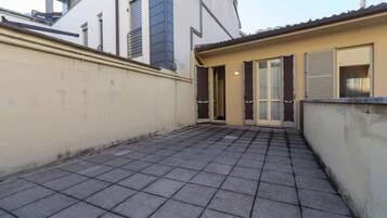 A courtyard with a tiled floor, a beige wall, and a building with a sloped roof and shutters.