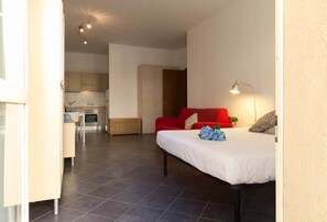 A compact living space with a bed, a red sofa, a small table, and a kitchenette in the background. - Galleria Roma Studio - Affitti Brevi Italia (Lecco)